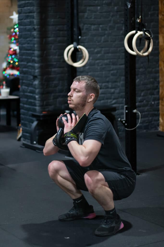 Un homme exécute un squat goblet, concentré et stable, tenant un kettlebell près de sa poitrine dans une salle de CrossFit décorée pour les fêtes, soulignant l'engagement envers la forme physique tout au long de l'année.