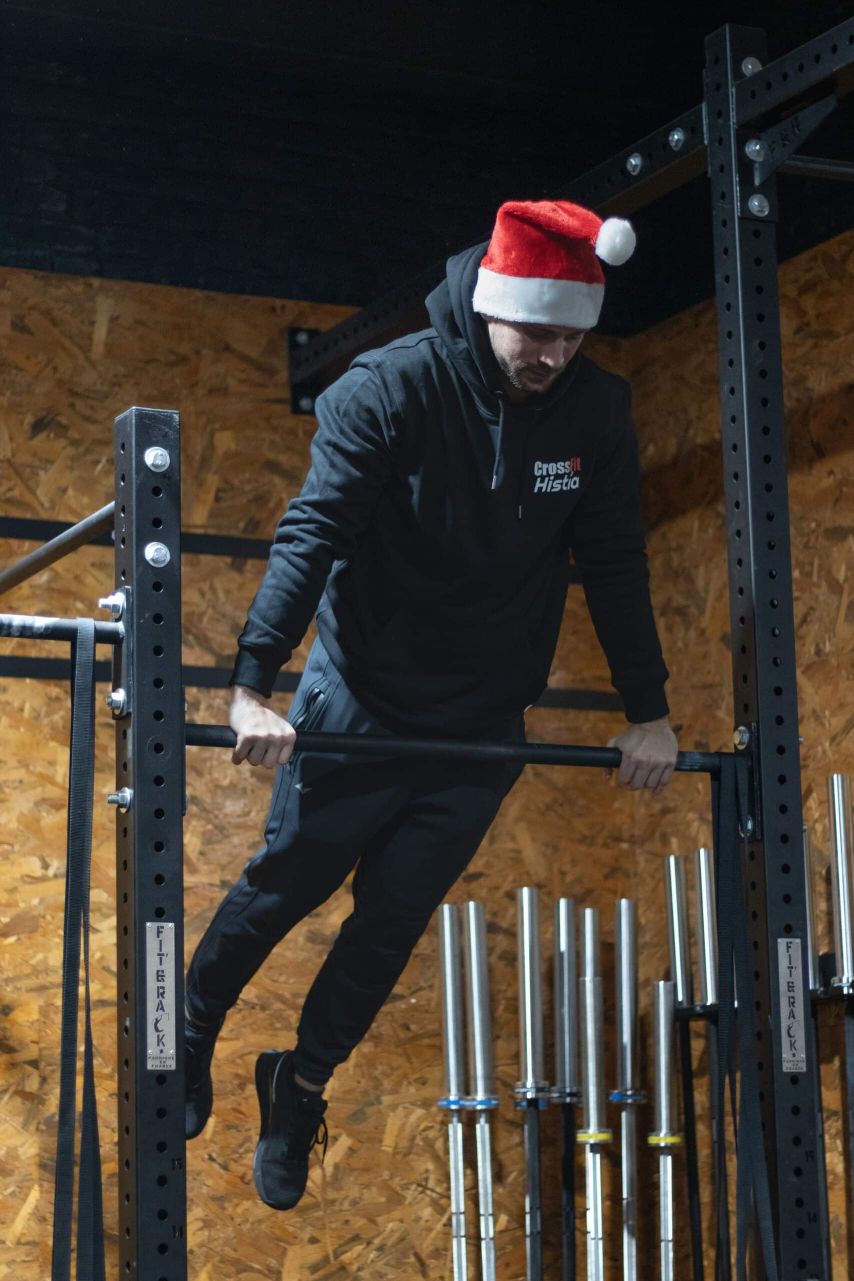 Coach de CrossFit Histía en tenue sportive et bonnet de Noël, réalisant un muscle-up sur une barre dans notre salle de sport à Valenciennes.